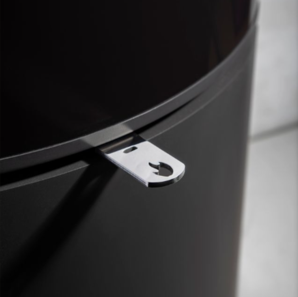 Close-up of black stovre with a small silver metal latch or tab attached near the rim, set against a softly blurred light gray background.