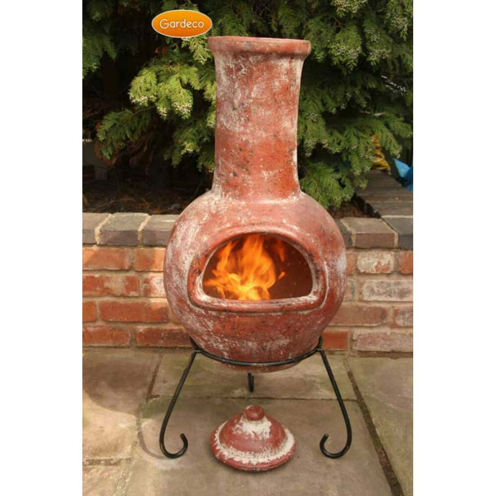 Terracotta chiminea with flames inside, set against a brick wall and greenery.
