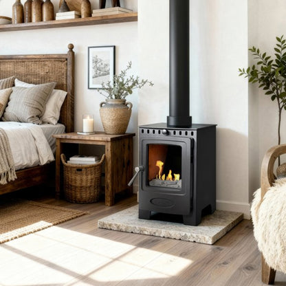 A black bioethanol stove with a visible fire, set on a stone hearth in a styled bedroom interior with a bed, side table, plants, and warm natural light.