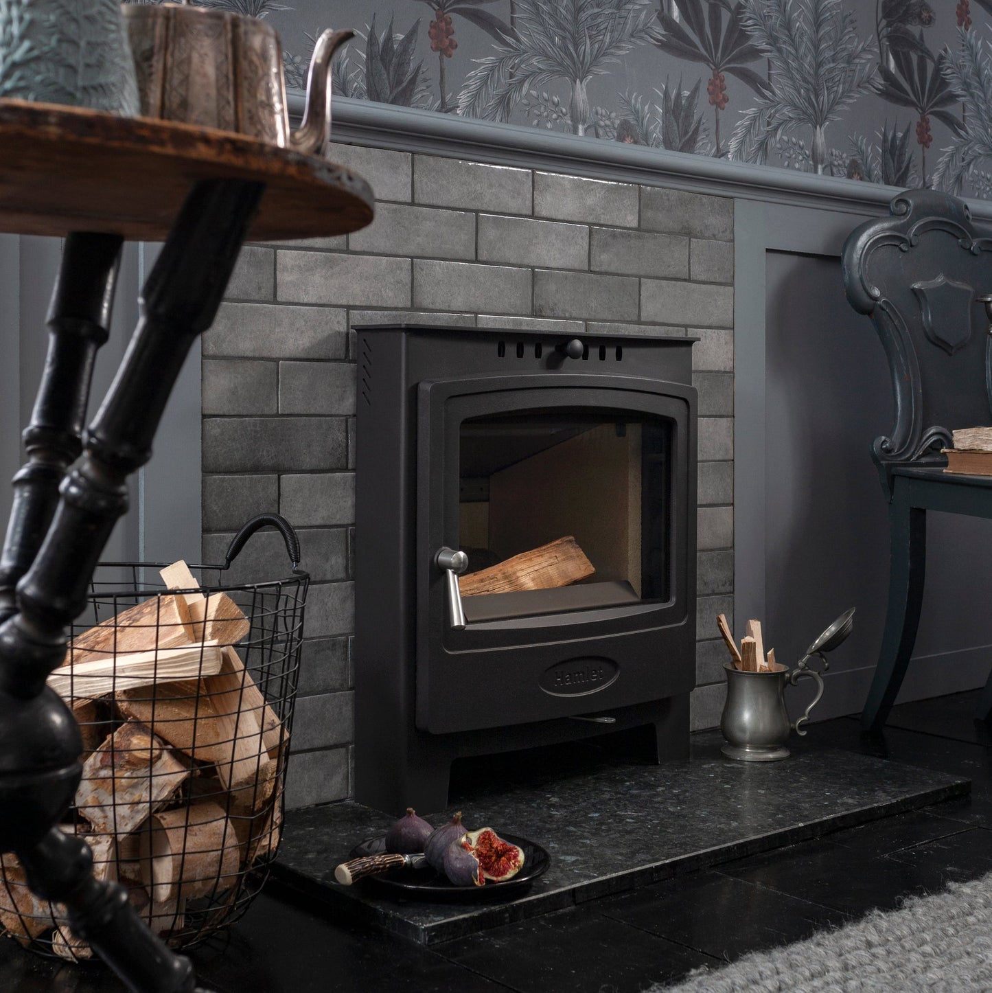 A black wood-burning stove set into a gray brick fireplace, styled in a cozy room with tropical wallpaper, a side table, and a gray rug.