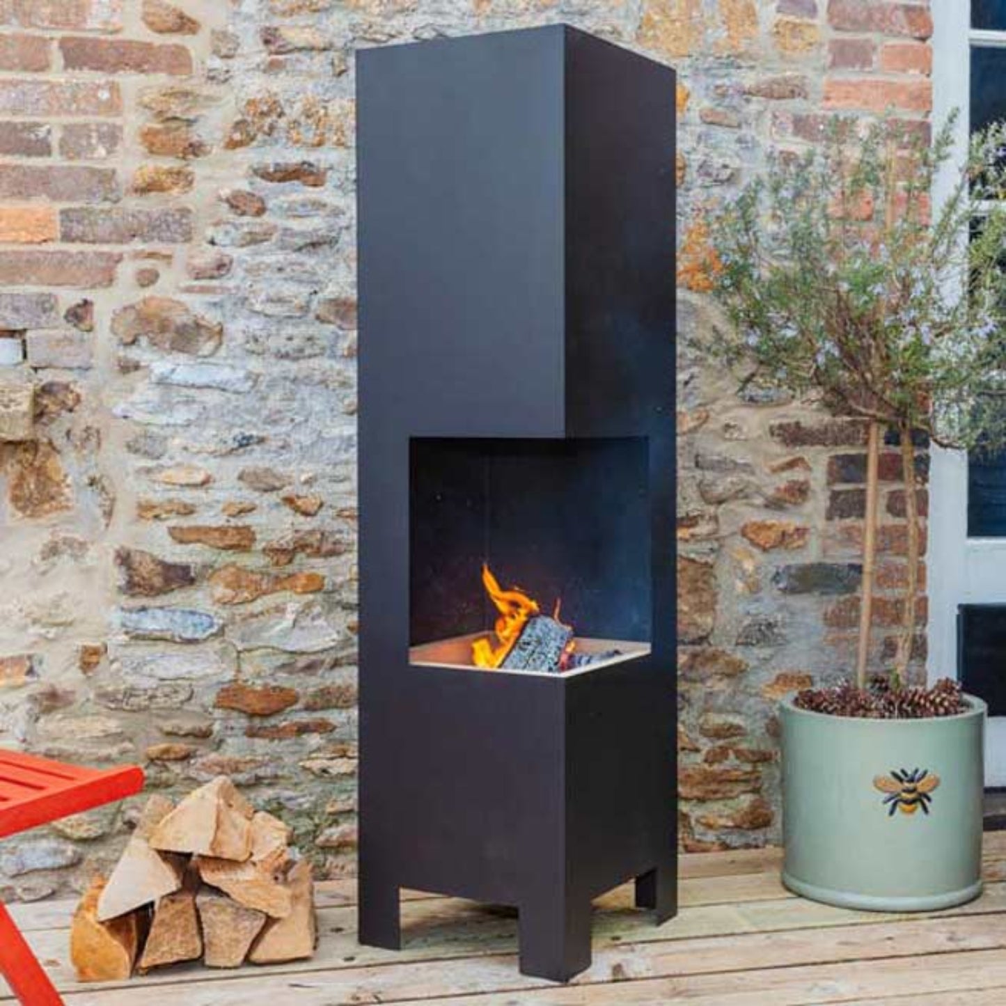 A tall, modern black outdoor fire pit with a visible flame, set on a wooden deck against a rustic stone wall, with a potted plant and stacked firewood nearby.