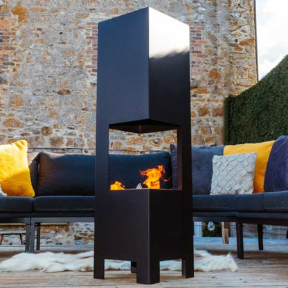 A modern black outdoor fire pit table with a visible flame, positioned in front of a dark outdoor sofa with yellow and gray pillows on a wooden deck against a stone wall background.