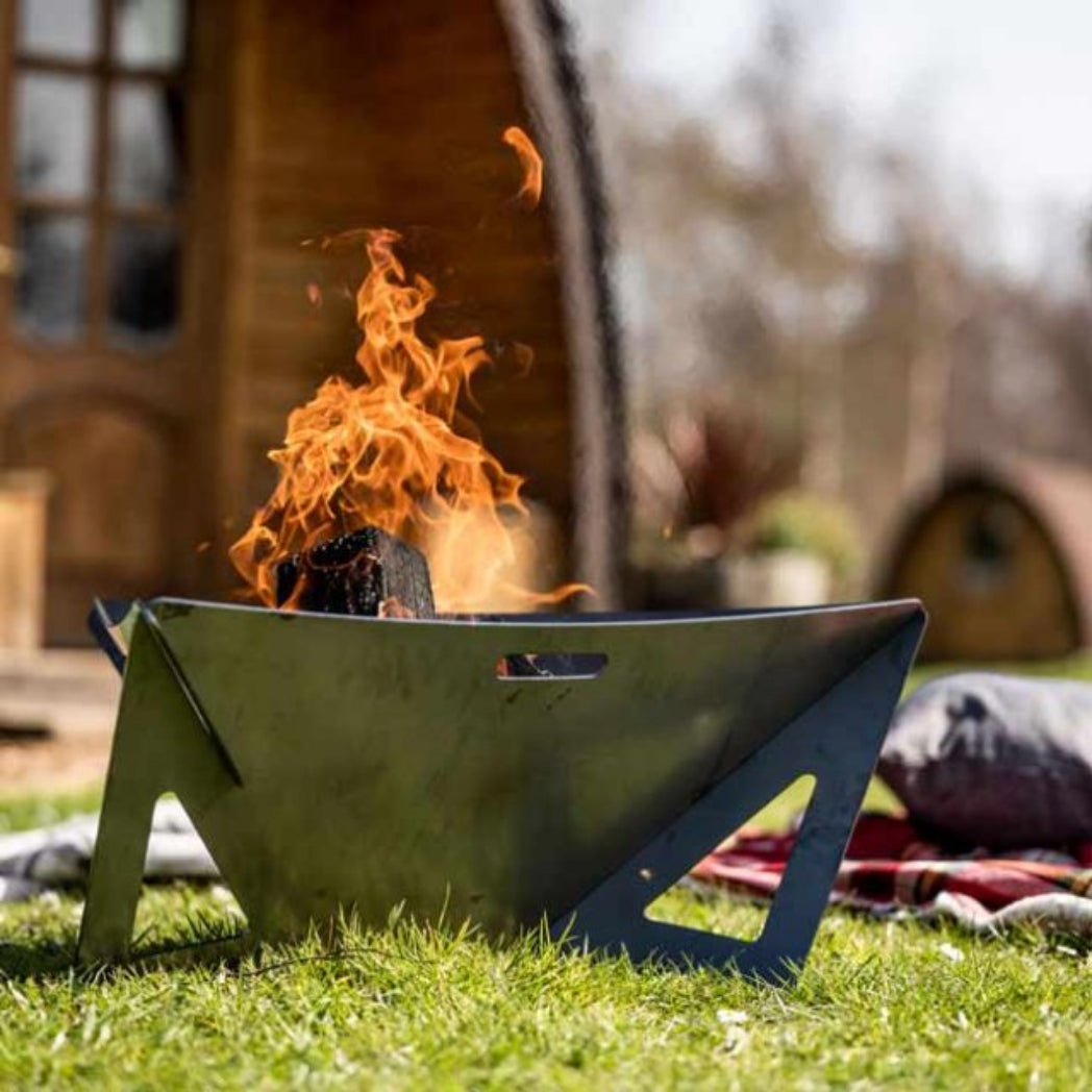 A metal campfire pit with a triangular design sits on green grass outdoors, with visible orange flames rising from inside. A blurred wooden cabin and another fire pit are in the background.