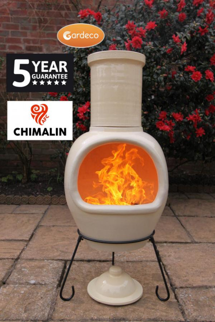 An off-white ceramic chiminea with a black metal stand, shown outdoors on a stone patio with a brick wall and red flowers in the background. The fire inside is lit with orange flames.