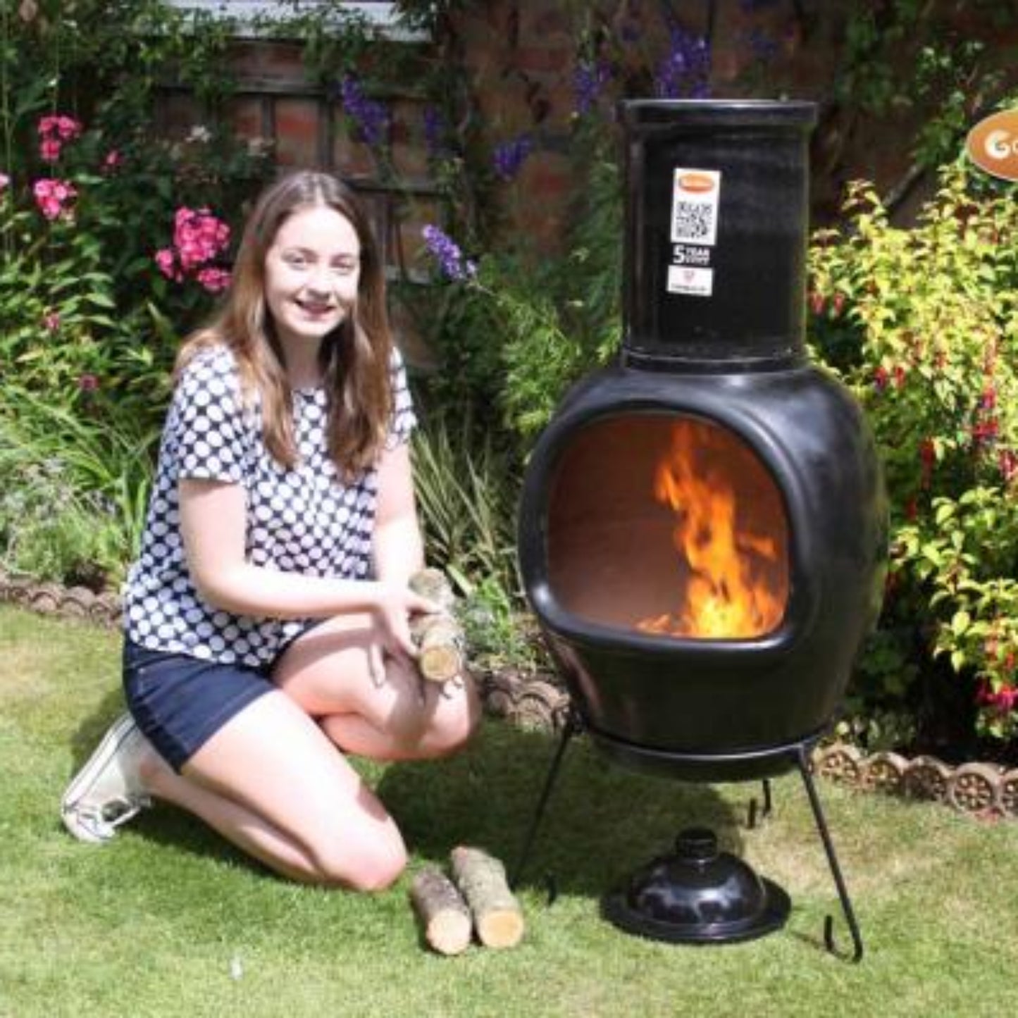 Woman kneeling next to a Chimalin chiminea in a garden with flowers and plants.