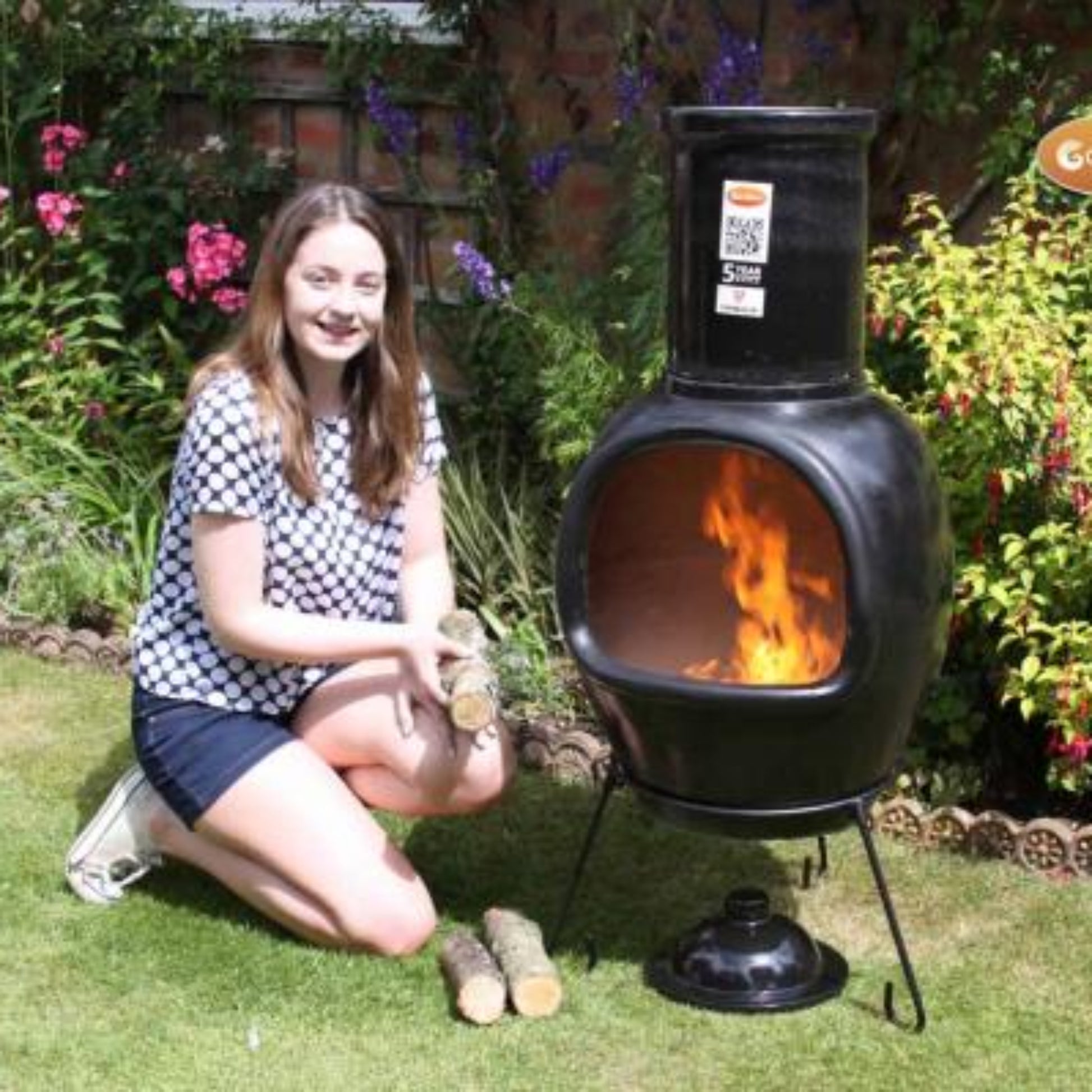 Woman kneeling next to a Chimalin chiminea in a garden with flowers and plants.