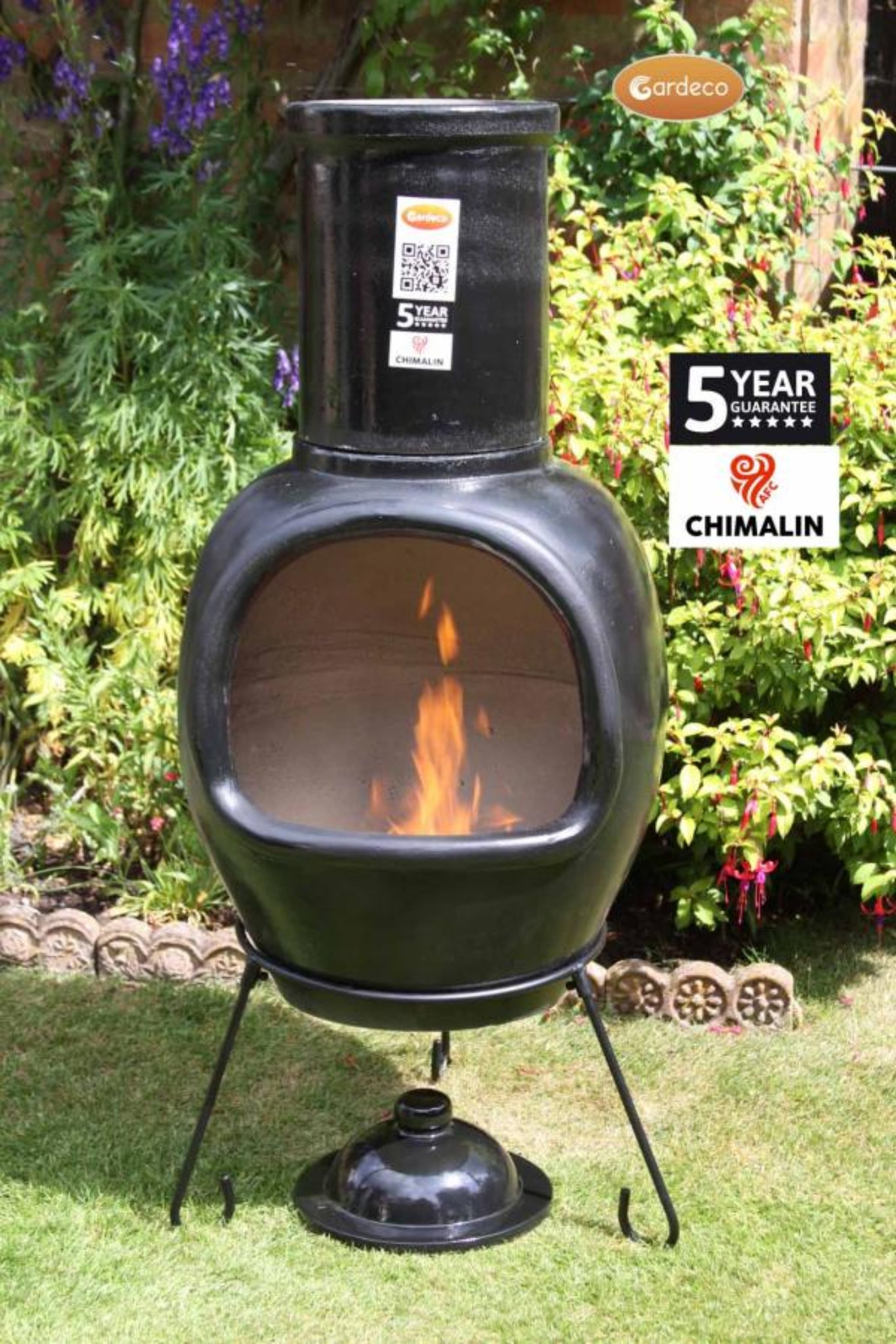 Black outdoor chiminea with flames on a grassy area with plants in the background, featuring Chimalin branding.
