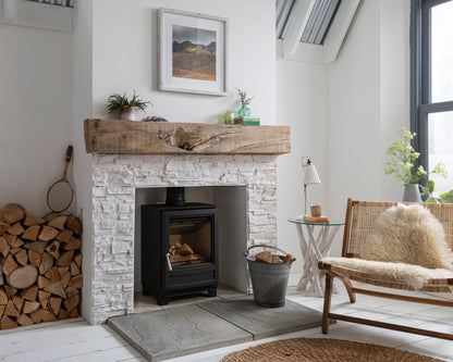 A black wood-burning stove installed in a white stone fireplace, styled in a bright, modern room with a rustic wooden mantel, stacked firewood, and a cozy woven chair.