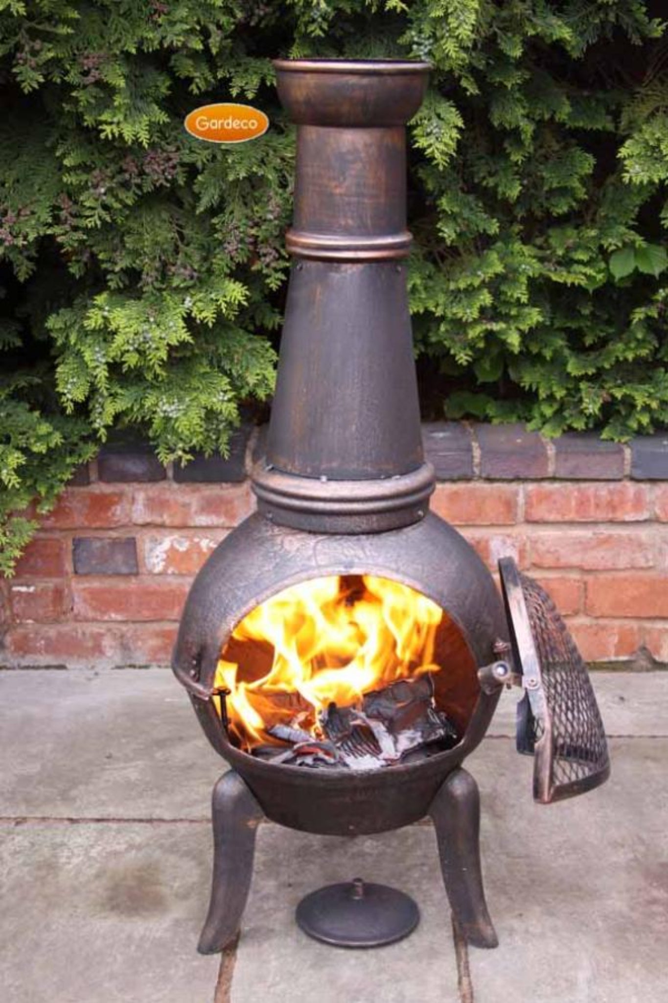 Cast iron chiminea with flames inside, surrounded by greenery and a brick wall.