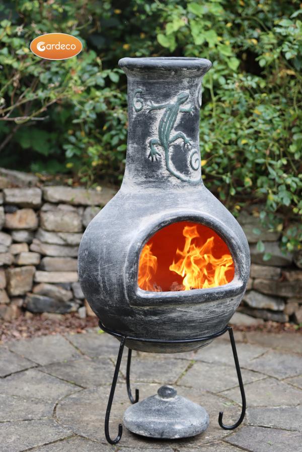 Outdoor chiminea with flames inside, set against a stone wall and greenery background.