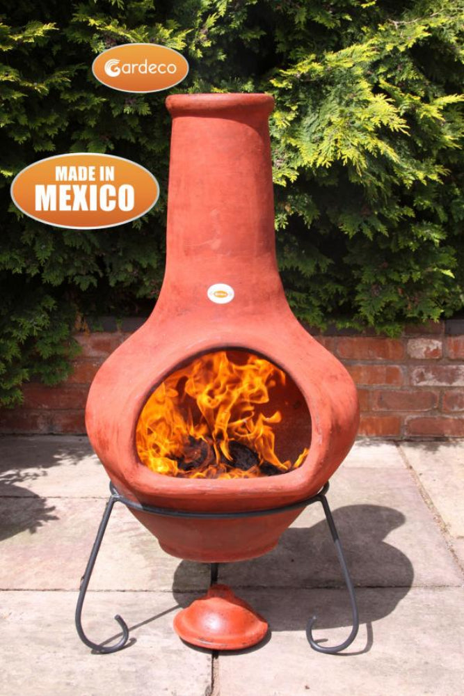 Terracotta chiminea with flames inside, branded 'Gardeco' and labeled 'Made in Mexico', against a green bush background.