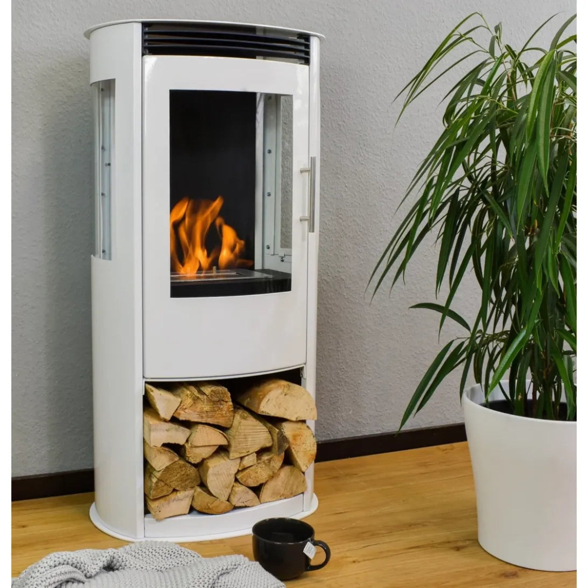 White freestanding fireplace with fire burning inside, logs below, and a plant to the right on a wooden floor.