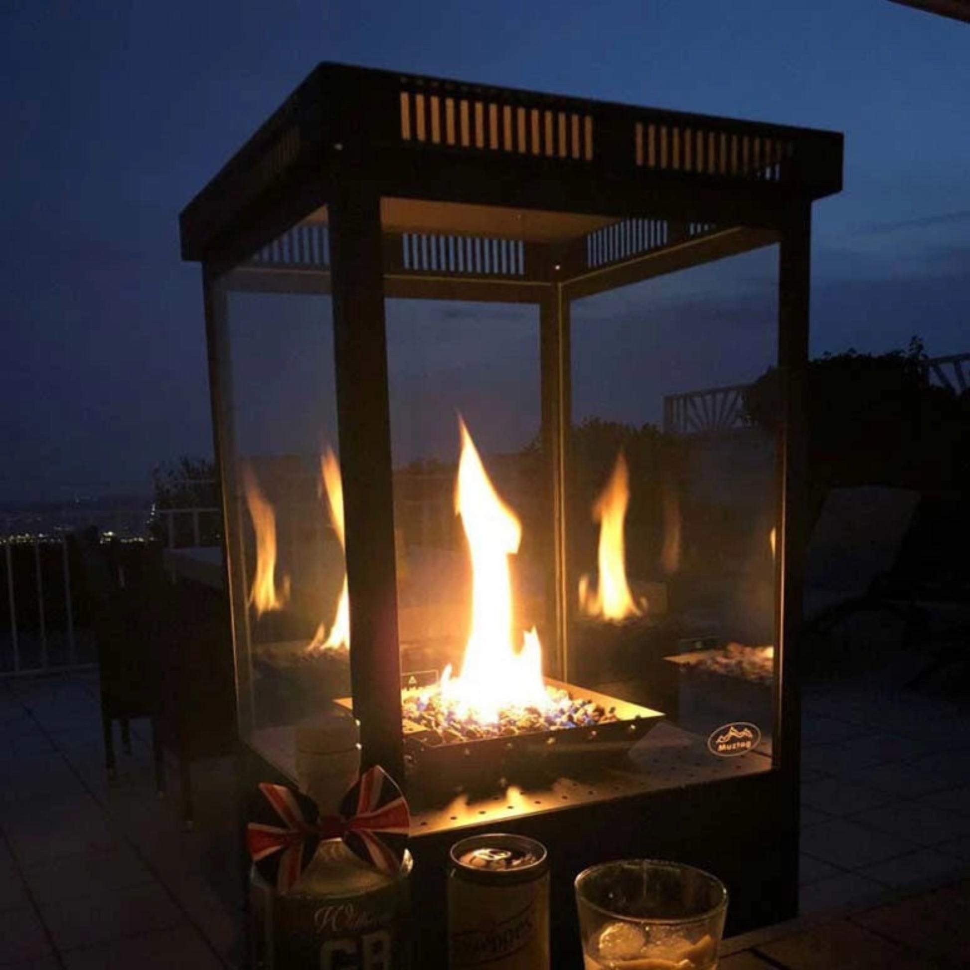 Outdoor fire pit with flames at night, surrounded by drinks and flags.