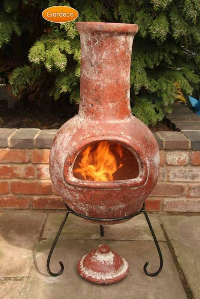 Terracotta chiminea with flames inside, set against a brick wall and greenery.