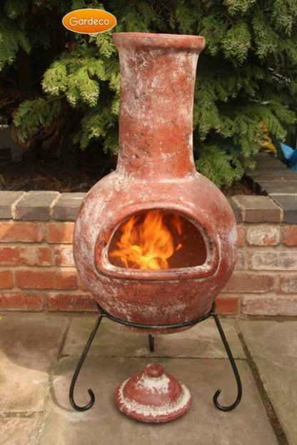 Terracotta chiminea with flames inside, set against a brick wall and greenery.