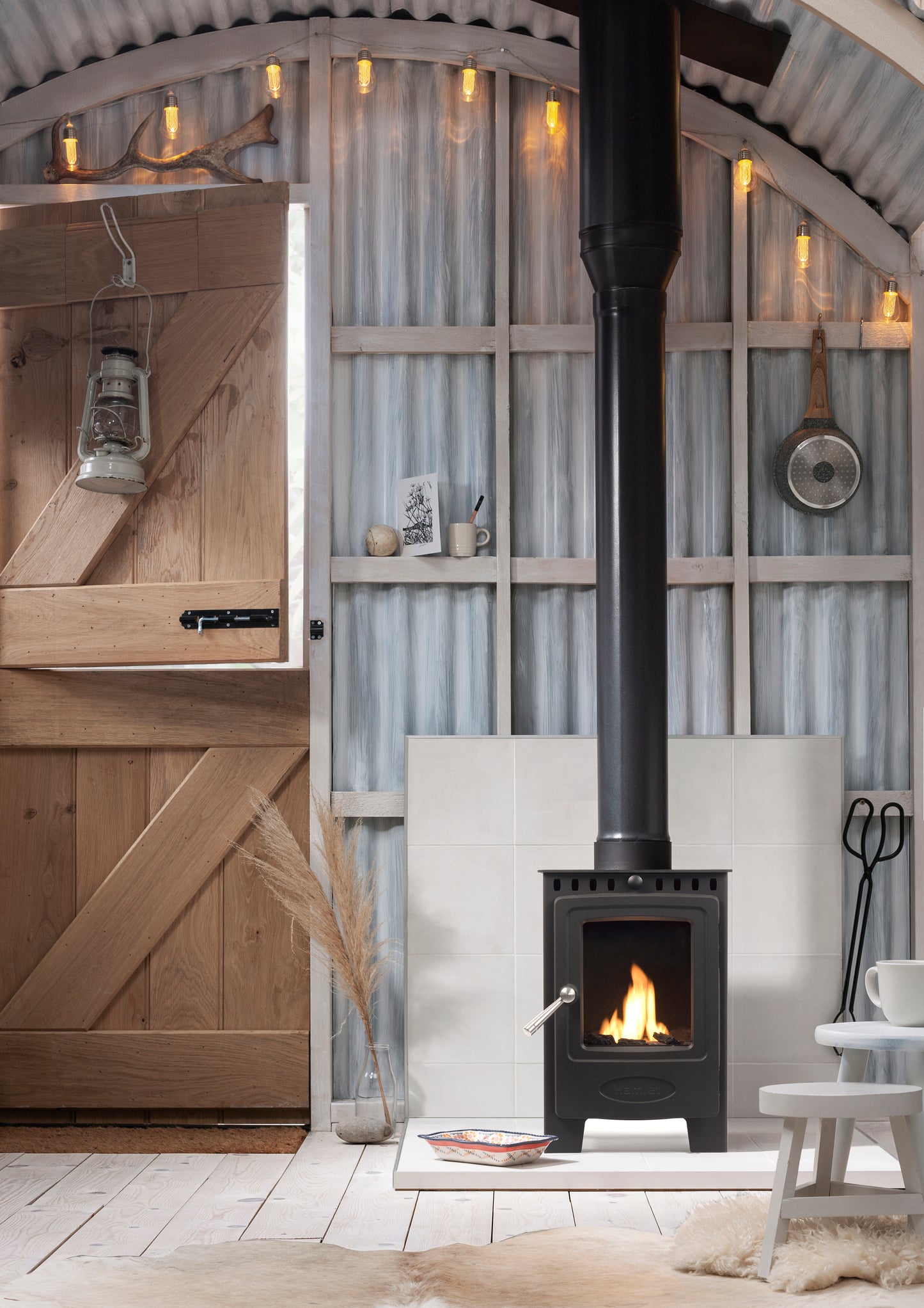 A black wood-burning stove with a visible fire, set in a rustic barn-style room with corrugated metal walls, wooden doors, and warm string lights.