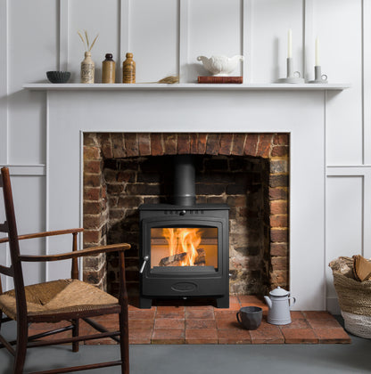 A black wood-burning stove with a visible fire, set into a brick fireplace within a white paneled wall. A white mantel holds decorative items, and a wooden chair and basket are visible in the room.