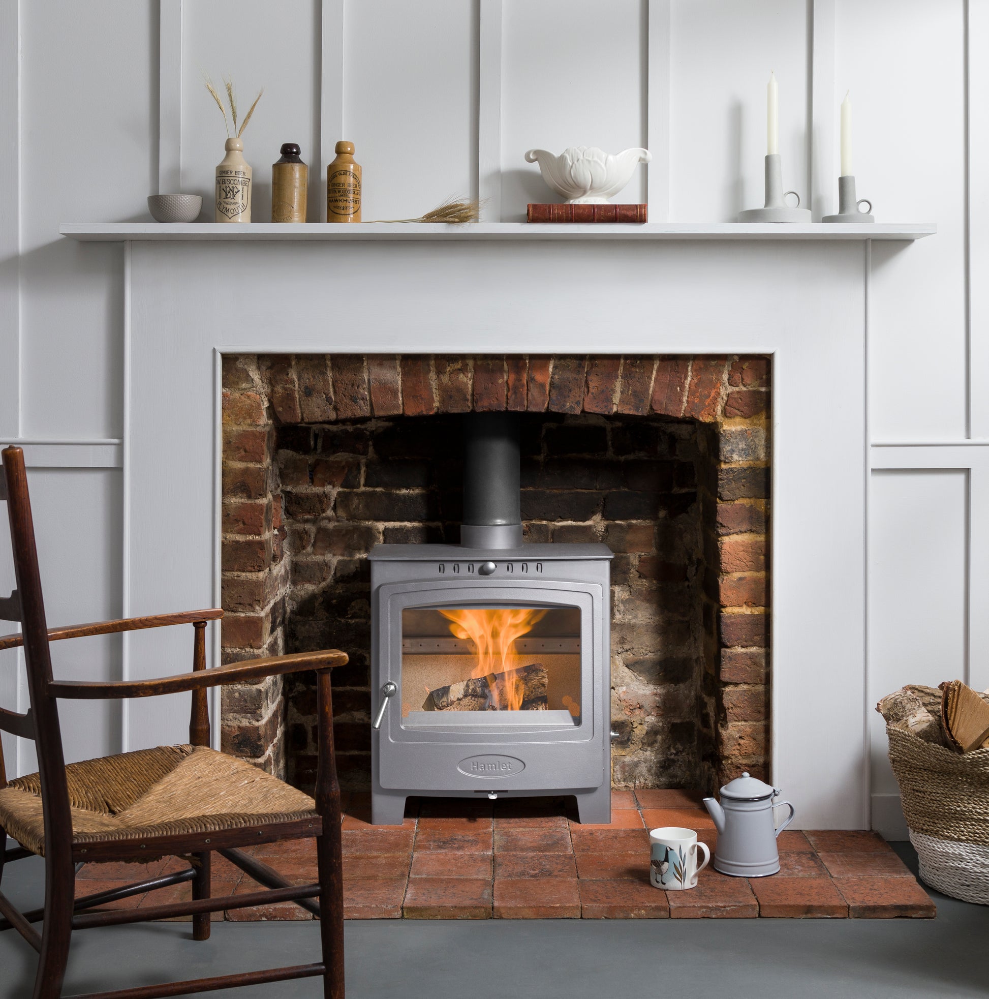 A silver wood-burning stove with a visible fire, set into a brick fireplace within a white paneled wall. A white mantel shelf with decorative items sits above, and a wooden chair and small table accessories are in the foreground.