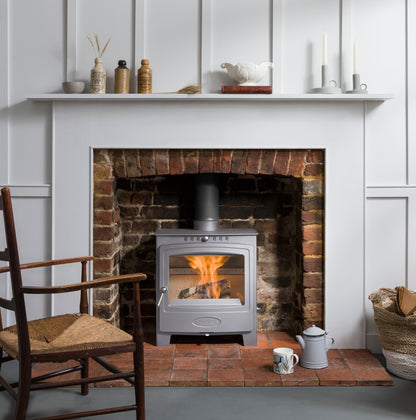 A silver wood-burning stove with a visible fire, set into a brick fireplace within a white paneled wall. A white mantel shelf with decorative items sits above, and a wooden chair and small table accessories are in the foreground.