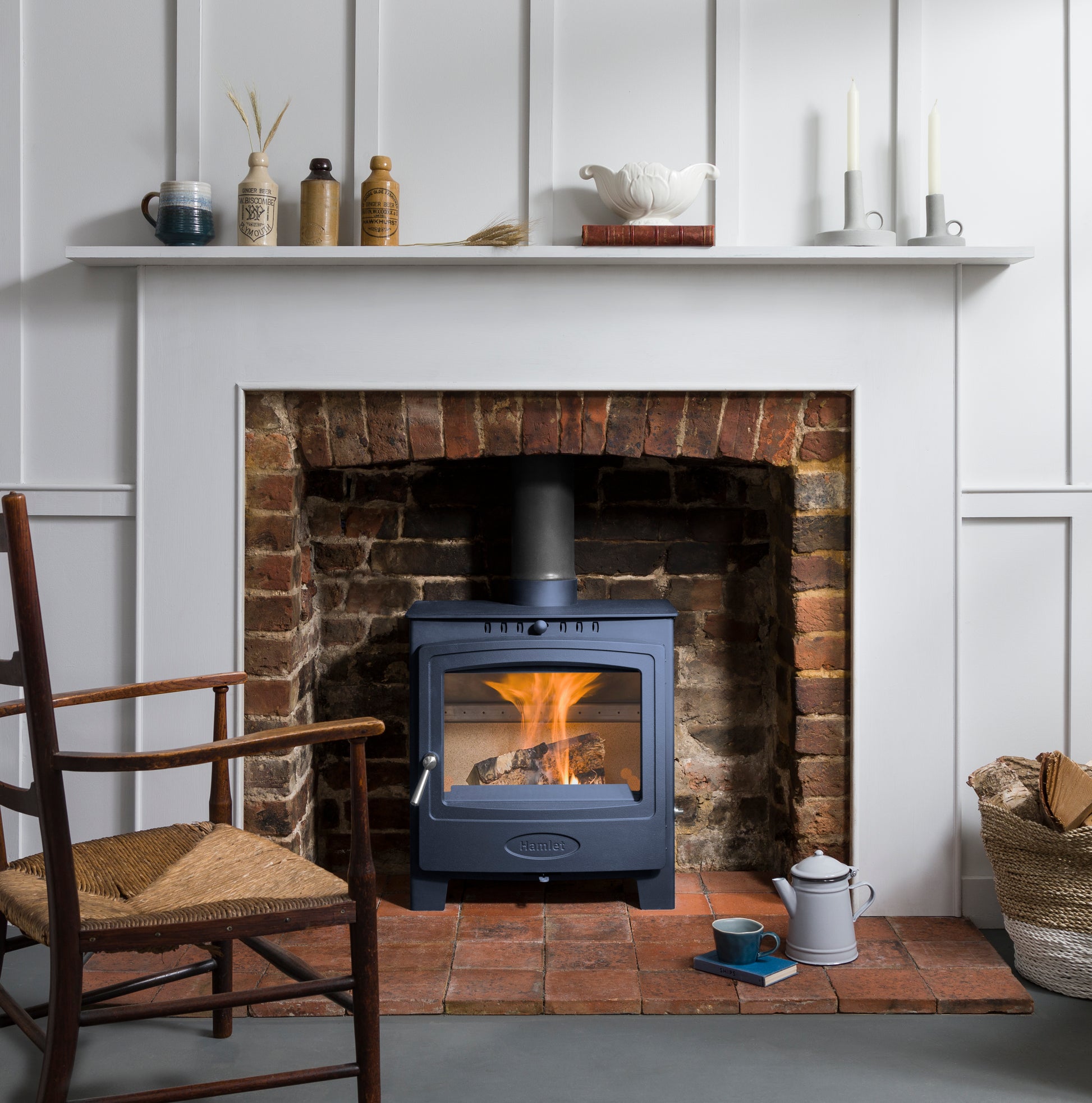 A dark blue wood-burning stove with a visible fire, set into a brick fireplace with white paneled walls, a white mantel with decorative items, a wooden chair, and a basket on the floor.