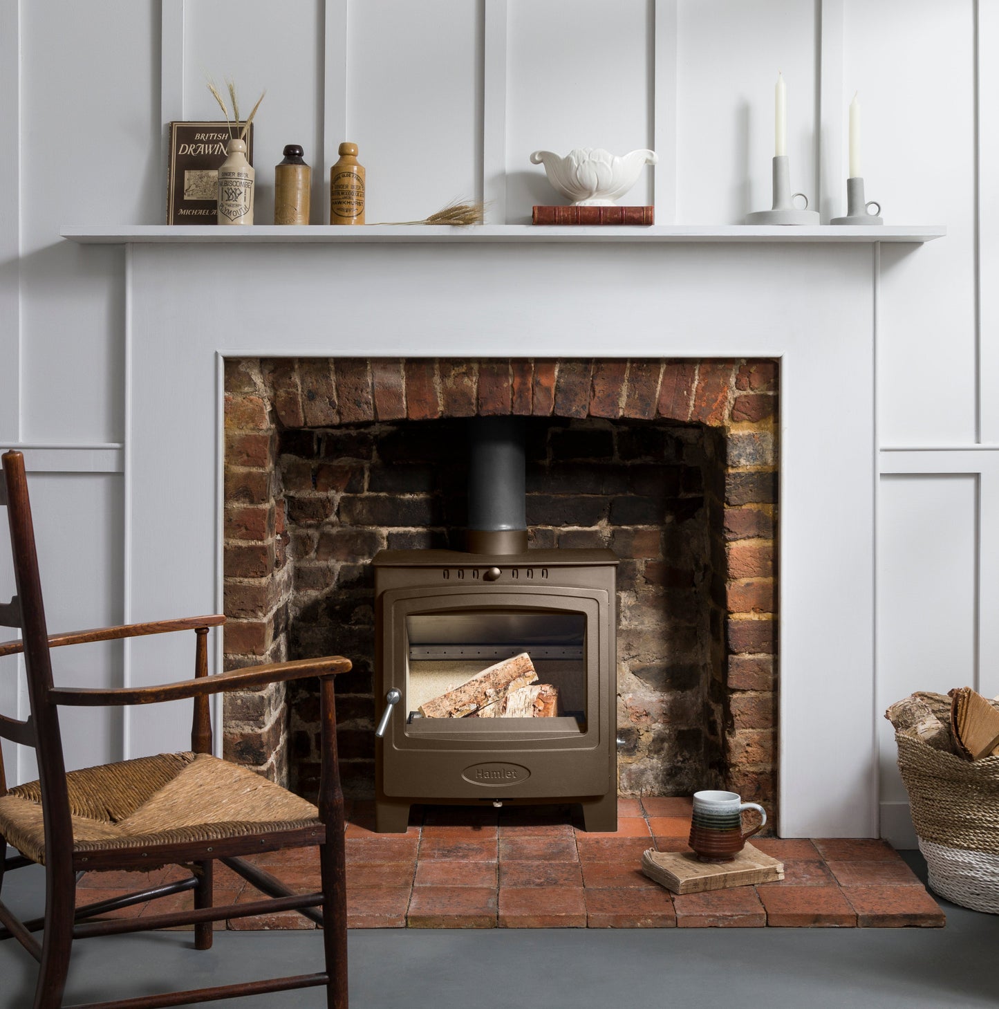 A bronze-brown wood-burning stove insert set into a brick fireplace with a white wall panel surround, styled in a cozy room with a chair, shelf decor, and a mug on the hearth.