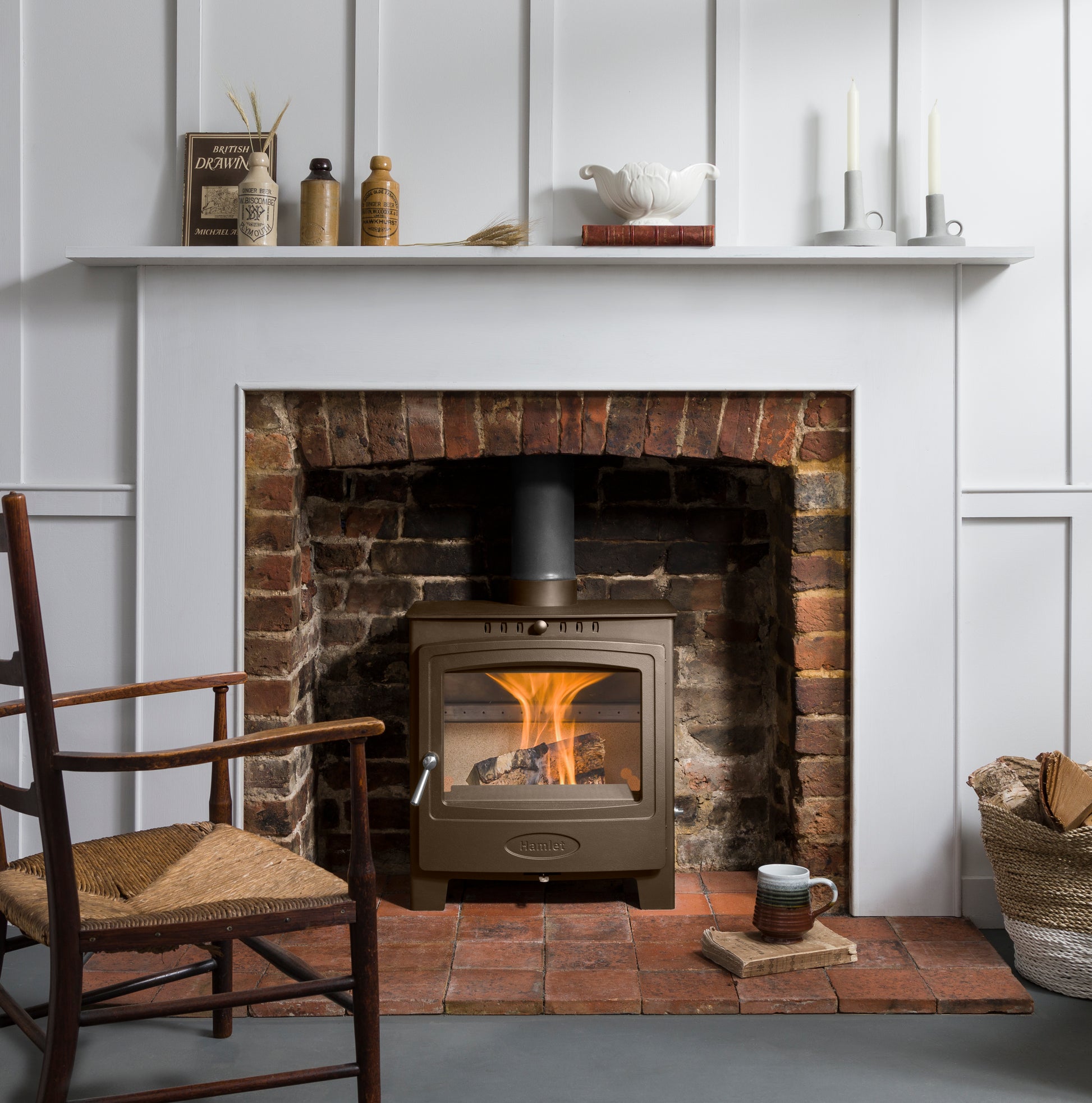 A bronze-brown wood-burning stove with a visible fire, set within a brick fireplace insert framed by a white wall panel. The scene is styled as a cozy interior with a white mantel holding decorative items, a wooden chair with a woven seat on the left, and a mug on the floor.