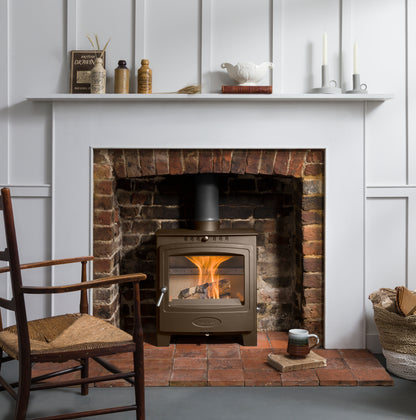 A bronze-brown wood-burning stove with a visible fire, set within a brick fireplace insert framed by a white wall panel. The scene is styled as a cozy interior with a white mantel holding decorative items, a wooden chair with a woven seat on the left, and a mug on the floor.