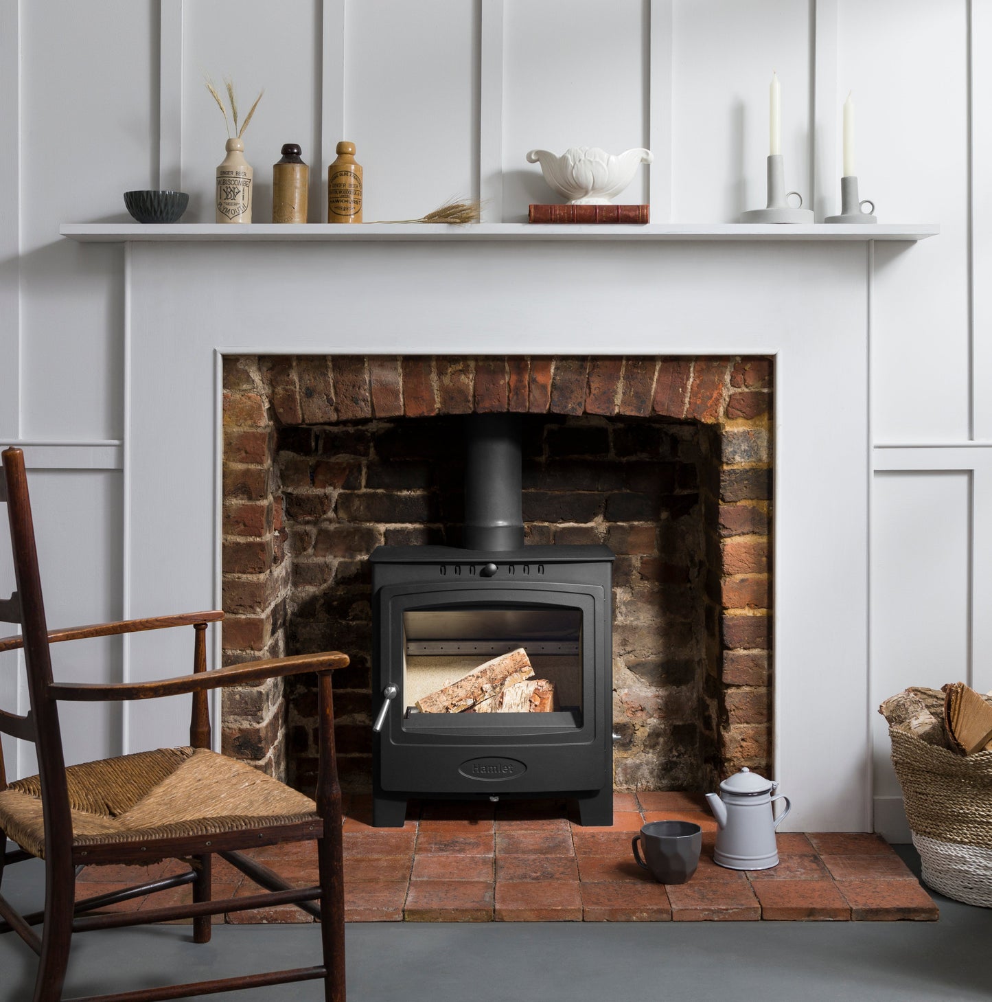 A black wood-burning stove with a glass door, set inside a rustic red brick fireplace. The fireplace is styled with a white mantel shelf holding decorative items, a wooden chair on the left, and a basket of firewood on the right.