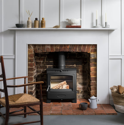 A black wood-burning stove with a glass door, set inside a rustic red brick fireplace. The fireplace is styled with a white mantel shelf holding decorative items, a wooden chair on the left, and a basket of firewood on the right.