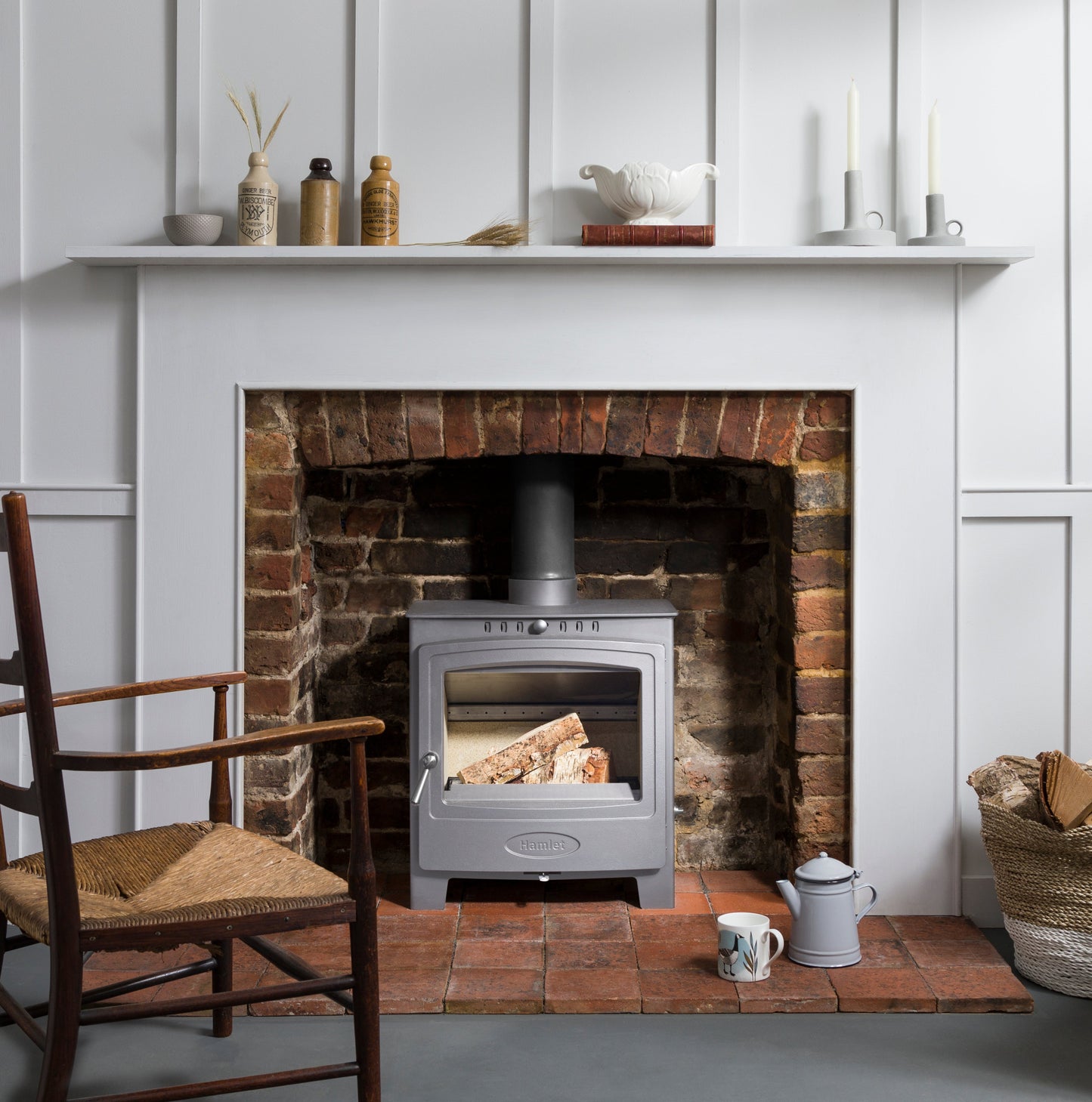 A silver wood-burning stove with a glass door sits inside a brick fireplace recessed in a white paneled wall. A white mantel shelf above holds decorative items, and a wooden chair and small table accessories are visible in the room.