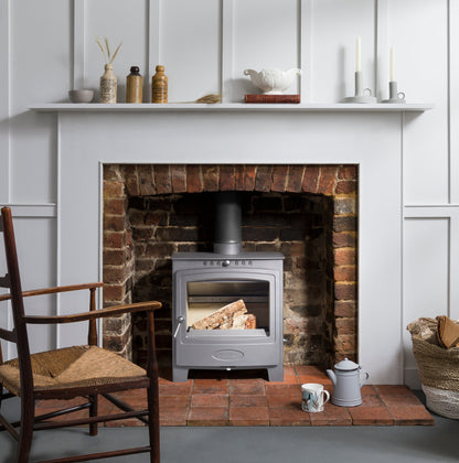 A silver wood-burning stove with a glass door sits inside a brick fireplace recessed in a white paneled wall. A white mantel shelf above holds decorative items, and a wooden chair and small table accessories are visible in the room.