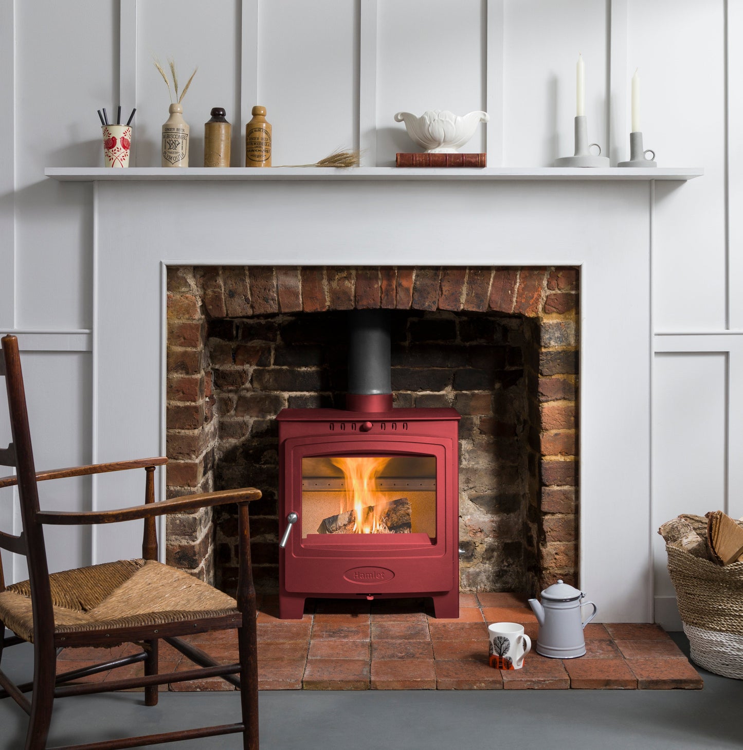 A red wood-burning stove with an active fire, set into a brick fireplace with a white mantel and wall paneling in the background.