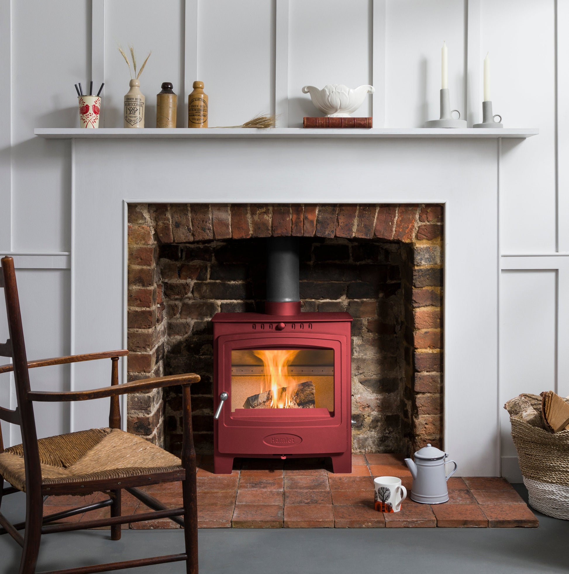 A red wood-burning stove with an active fire, set into a brick fireplace with a white mantel and wall paneling in the background.
