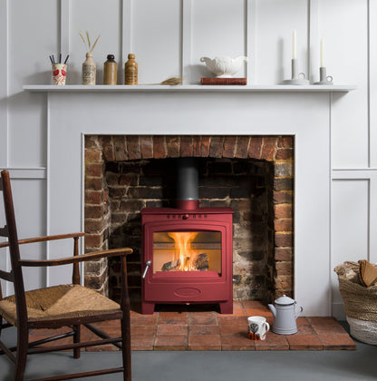 A red wood-burning stove with an active fire, set into a brick fireplace with a white mantel and wall paneling in the background.