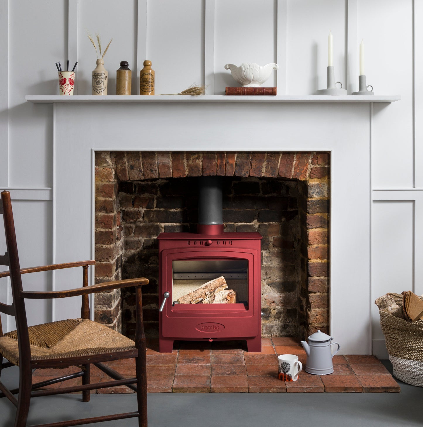 A red wood-burning stove with an active fire, set into a brick fireplace with a white mantel and wall paneling in the background.
