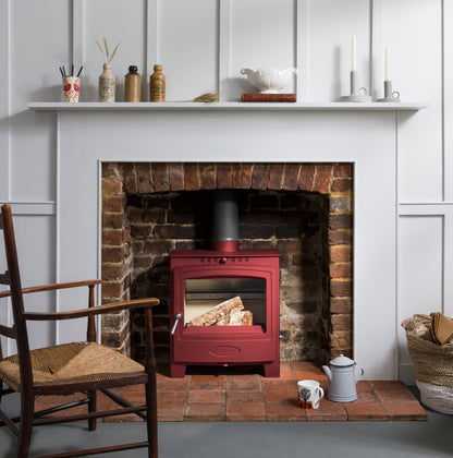 A red wood-burning stove with an active fire, set into a brick fireplace with a white mantel and wall paneling in the background.