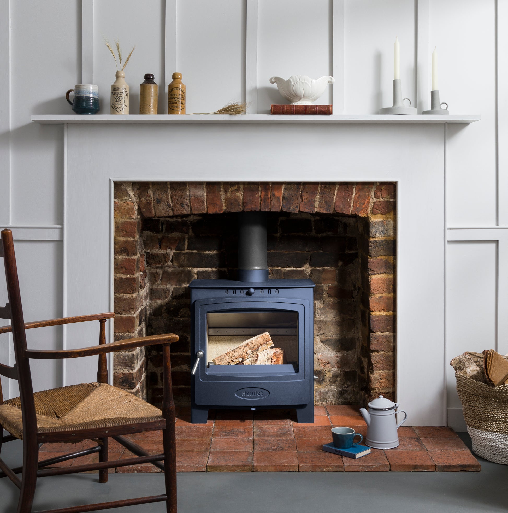 A dark blue wood-burning stove with a glass door, set inside a rustic red brick fireplace. The fireplace is styled with a white mantel, decorative ceramic vases, candles, and a woven basket chair on the left.