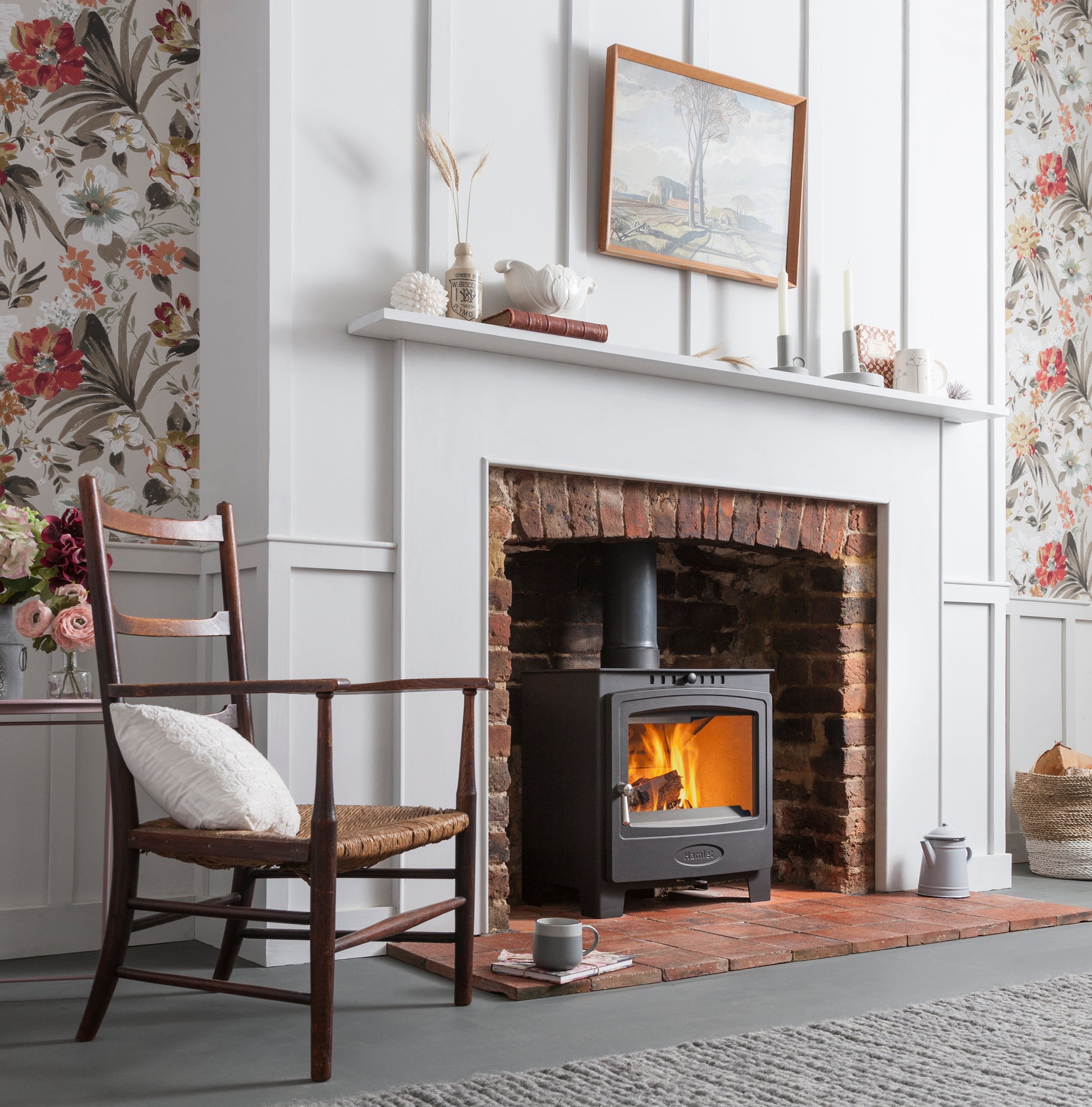 A black wood-burning stove with a glowing fire, set into a white brick fireplace in a styled room with floral wallpaper, a wooden chair, and a white mantel.