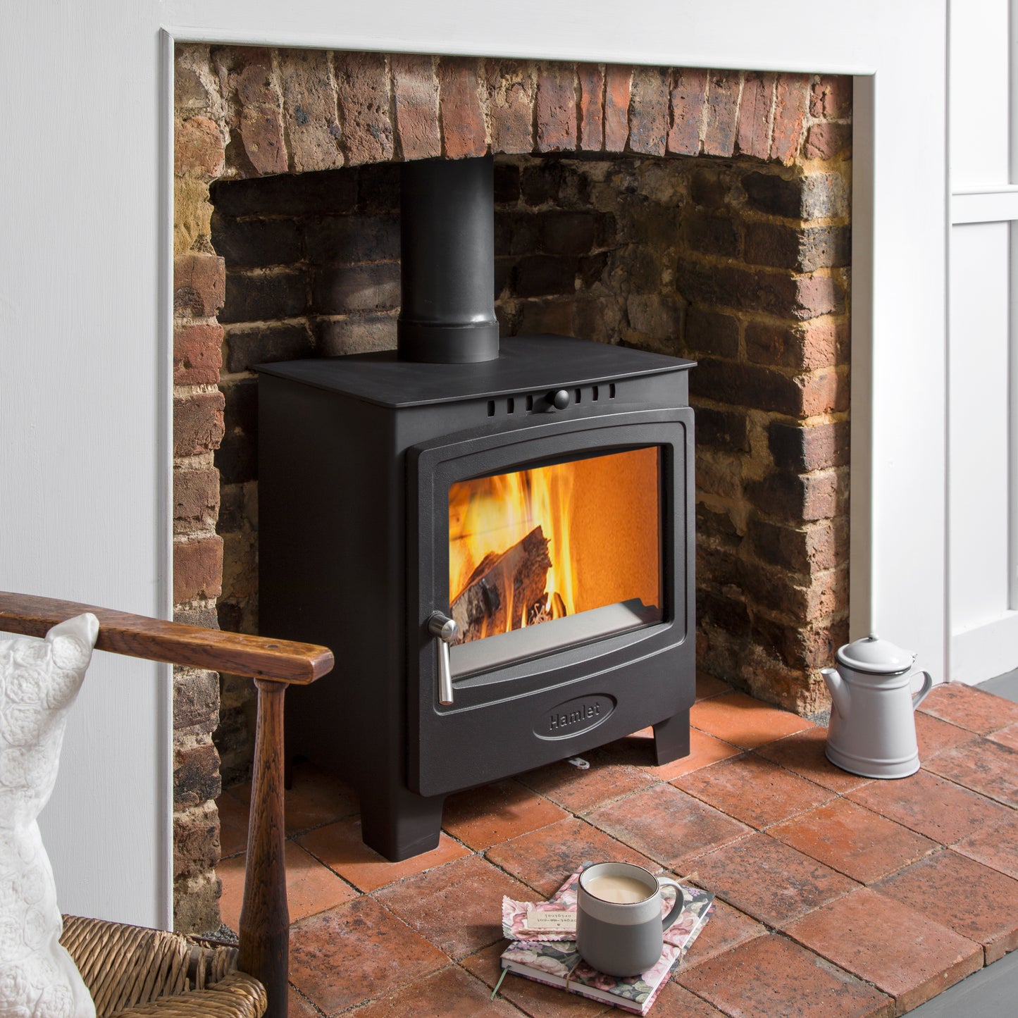 A black wood-burning stove with a visible fire, set against a brick fireplace in a styled room scene with a mug and teapot on the floor.