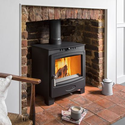 A black wood-burning stove with a visible fire, set against a brick fireplace in a styled room scene with a mug and teapot on the floor.