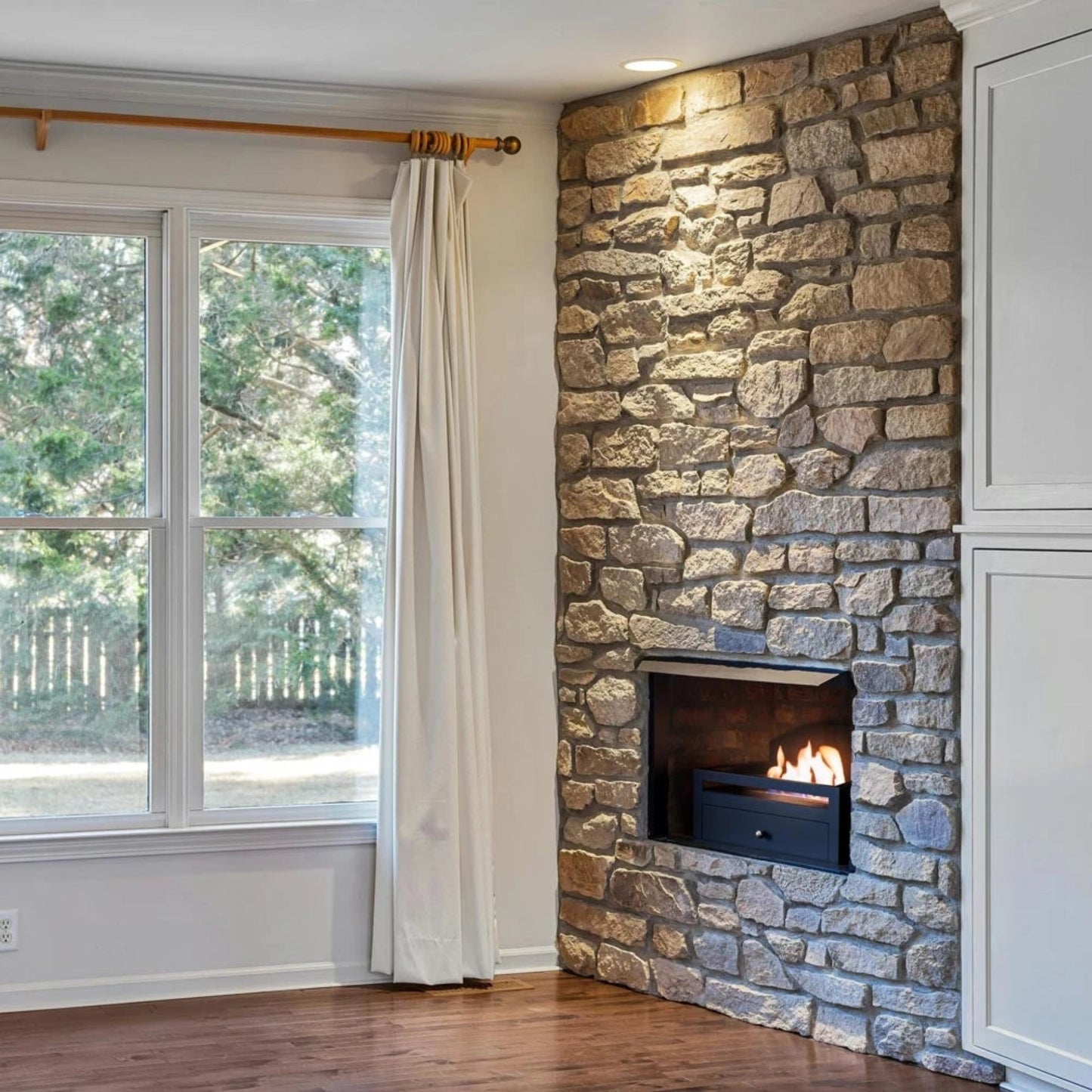 Scandiflames Kingston Freestanding Bioethanol Fireplace Insert in a stone fireplace in a room with a large window and white curtains.