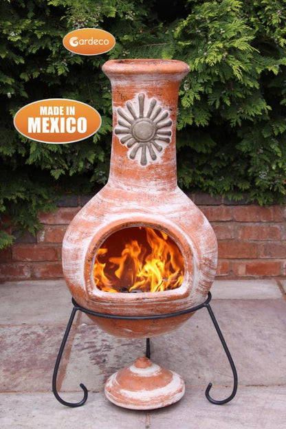 Terracotta chiminea with flames inside, made in Mexico, against a green bush background.