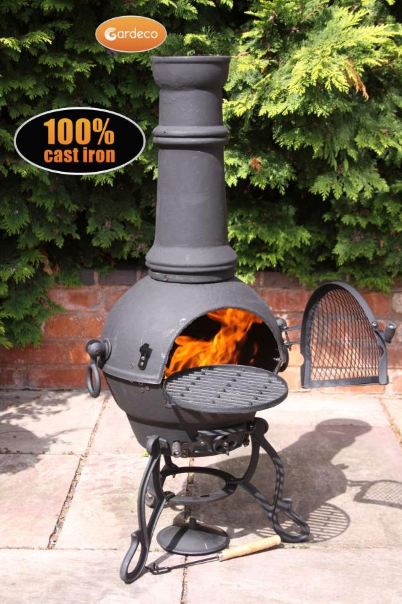 Cast iron chiminea with flames inside, surrounded by greenery and a brick wall.