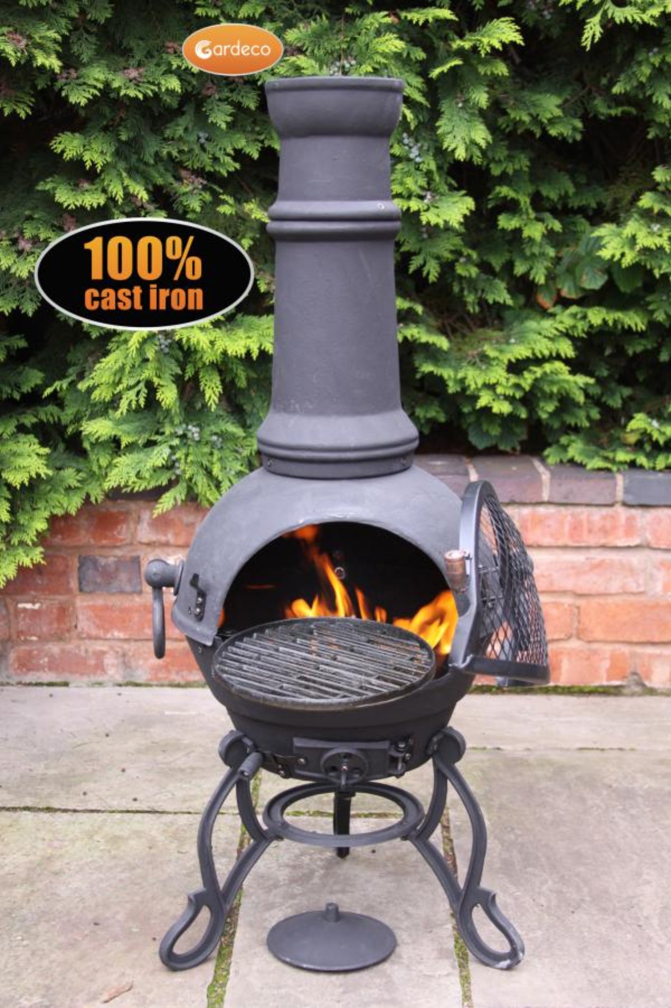 Cast iron chiminea with flames inside, surrounded by greenery and a brick wall.