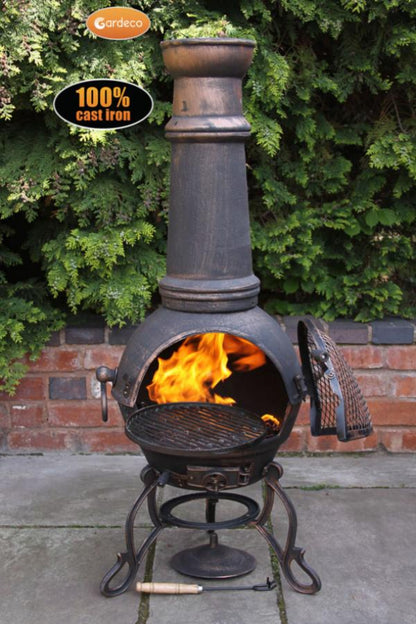 Cast iron chiminea with flames inside, surrounded by greenery and a brick wall.