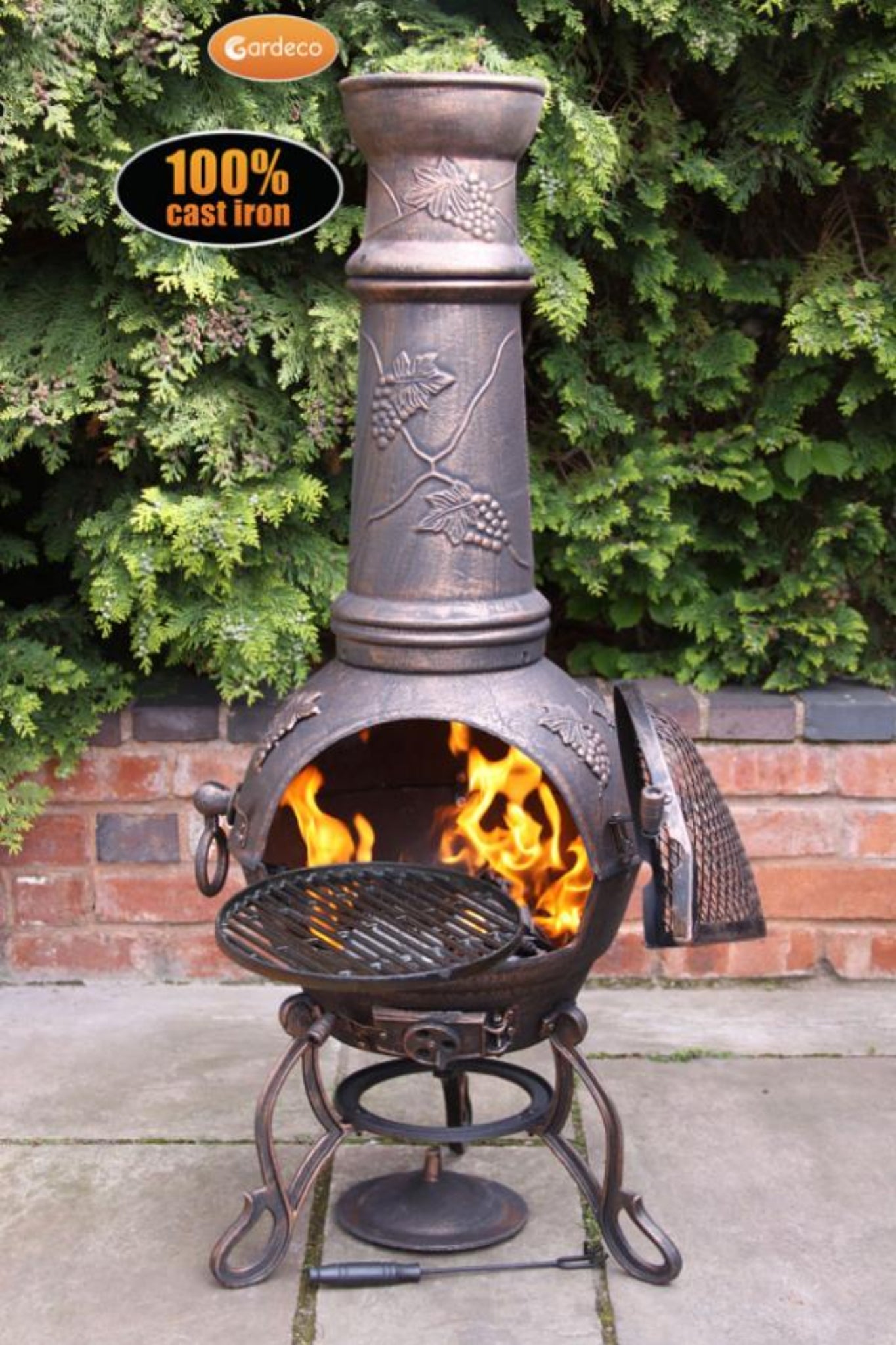 Cast iron chiminea with flames inside, surrounded by greenery and a brick wall.