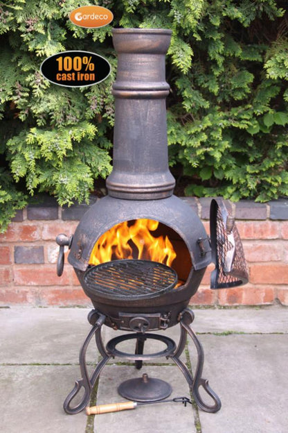 Cast iron chiminea with flames inside, surrounded by greenery and a brick wall.