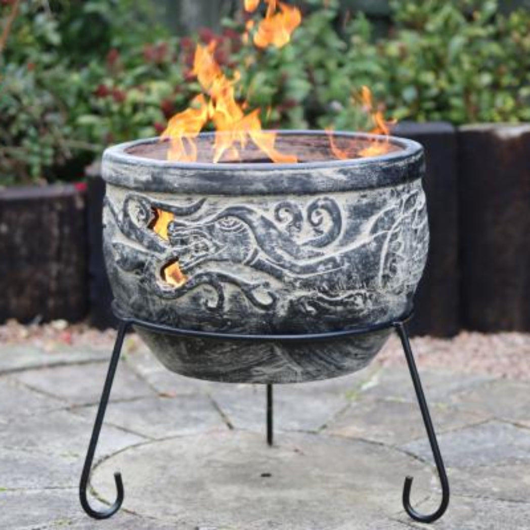 A round outdoor fire pit with a dark gray stone-look body featuring embossed scrollwork, supported by a black metal stand with curved legs. Flames are burning inside the fire pit, set on a stone patio with a wooden fence and green garden foliage in the background.