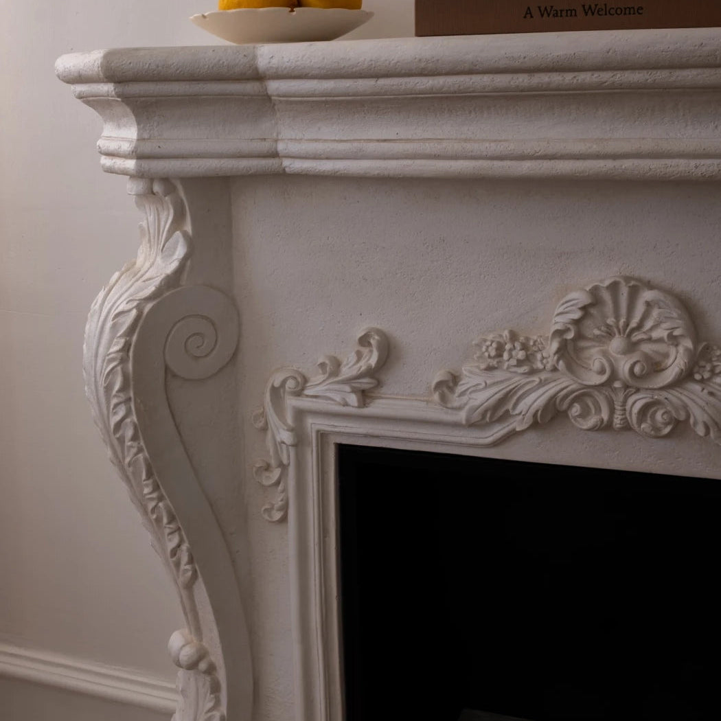 Decorative white fireplace with ornate details, a bowl of oranges, and books on a plain wall.
