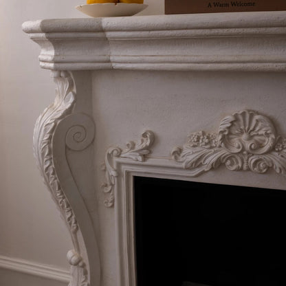 Decorative white fireplace with ornate details, a bowl of oranges, and books on a plain wall.